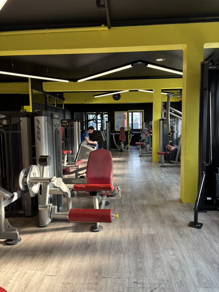 Salle de musculation moderne équipée de machines BH Fitness avec murs jaunes, sol en bois et éclairage LED design.